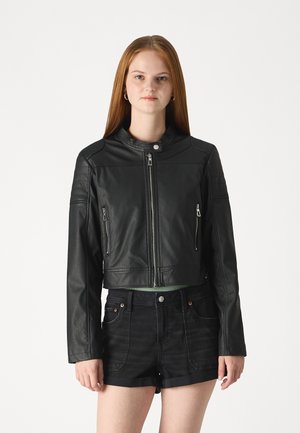 Veste en cuir noir courte avec deux poches zippées, un col à pression et des accents texturés sur les épaules. Associée à un short noir.
