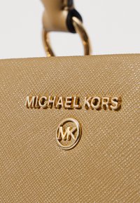Złota torebka z chropowatej skóry z tłoczonym logo "MICHAEL KORS" oraz okrągłym emblematem "MK", z gładkim czarnym uchwytem akcentującym.