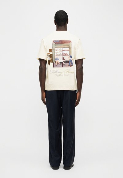 Άνδρας που φοράει κρεμ μπλουζάκι με σκηνή αρτοποιείου και κείμενο «Filling Pieces Breaking Bread» στην πλάτη, σε συνδυασμό με σκούρο παντελόνι με λεπτές ρίγες.