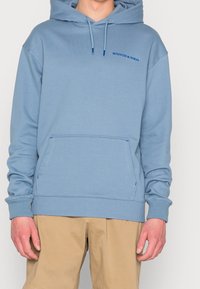 Sudadera de color azul claro hecha de una tela suave, con un bolsillo tipo canguro, puños y dobladillo acanalados. Incluye el logo "SCOTCH & SODA" en un azul más oscuro.