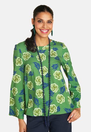 Eine lächelnde Frau trägt eine grüne Bluse mit Blumenmuster und eine blaue Halskette. Sie posiert selbstbewusst vor einem weißen Hintergrund. Ihr dunkles Haar ist elegant frisiert.
