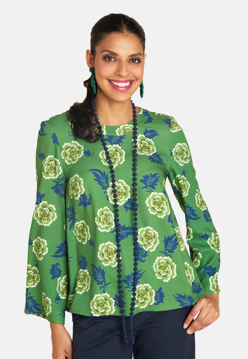 Eine lächelnde Frau trägt eine grüne Bluse mit Blumenmuster und eine blaue Halskette. Sie posiert selbstbewusst vor einem weißen Hintergrund. Ihr dunkles Haar ist elegant frisiert.