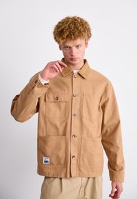 Beige bomullsjakke med krage, to brystlommer og to nedre lommer. Sølv trykknapper og en blå etikett som akcent.