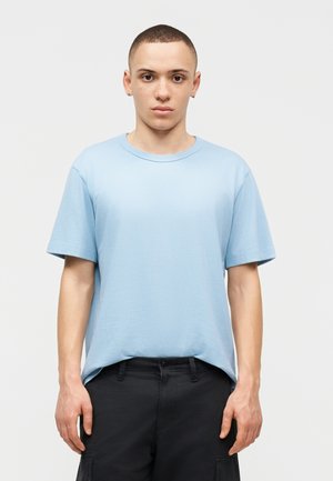 Νεαρός με κοντά μαλλιά, που φοράει γαλάζιο μπλουζάκι και μαύρο παντελόνι, στέκεται μπροστά σε απλό ανοιχτόχρωμο φόντο, κοιτώντας μπροστά.