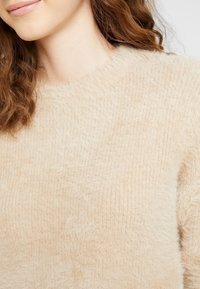 Beige tröja med en texturerad, fluffig stickning och en rundad halsringning, som visar upp ett mjukt, fluffigt tyg och subtila variationer i färg.