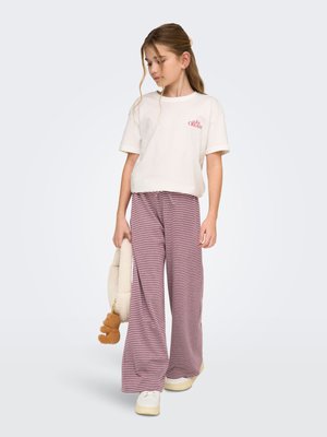 Dziewczyna w białej koszulce z czerwonym napisem, szerokich wąskich spodniach w paski, białych trampkach, trzymająca pluszową torebkę z breloczkiem w formie pompona, patrząca na bok.