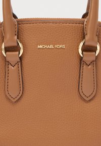 Καφέ τσάντα χειρός από δέρμα με υφή, με εξαρτήματα σε χρυσό τόνο, διπλές λαβές και ένα χρυσό λογότυπο με επωνυμία. Ομαλές ραφές βελτιώνουν το σχεδιασμό.