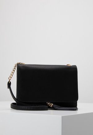 Zwarte leren crossbodytas met gouden kettingriem en tassel-detail, geplaatst op een wit oppervlak tegen een lichtgrijze achtergrond.