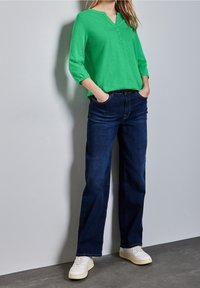 Grön långärmad topp med henley-halsringning, i kombination med mörkblå vida jeans och vita sneakers på en texturerad grå golv.