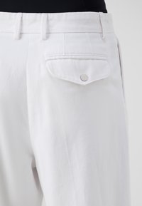 Pantalones de algodón blancos con un frente plano, bolsillos estándar y un bolsillo trasero único con un detalle de botón plateado. Textura suave.