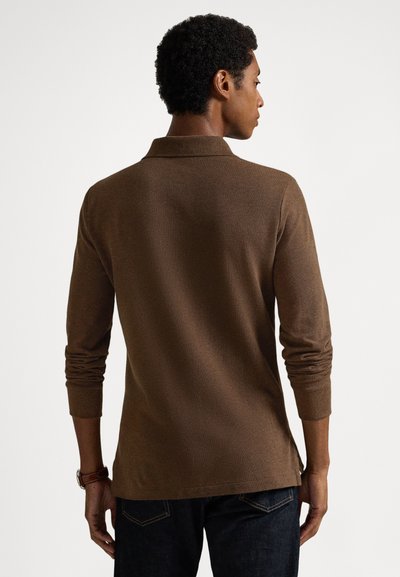 Brūna garroku polokrekls ar teksturētu adījumu, salocītu apkakli un sānu šķēlumiem, kombinēts ar tumšām džinsa biksēm.