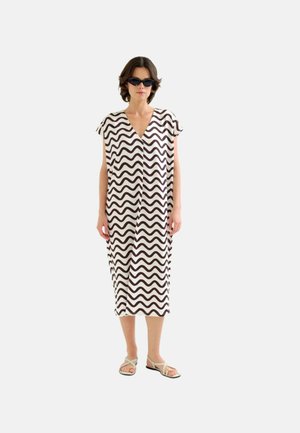 Mujer con vestido midi de rayas onduladas en blanco y negro, gafas de sol negras y sandalias beige, de pie sobre un fondo claro y liso.