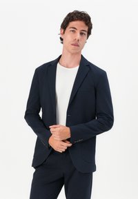Blazer azul-marinho feito de tecido suave, design de um botão, corte ajustado, com um bolso no peito e punhos com botão, combinado com uma camisa branca.