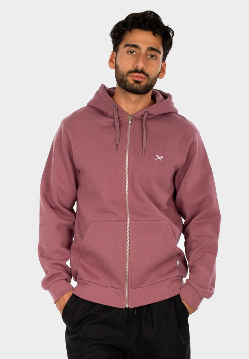 Mann mit dunklem Haar und Bart, der einen mauvefarbigen Hoodie mit Reißverschluss trägt und die Hände in den Taschen hat, vor einem schlichten hellen Hintergrund.