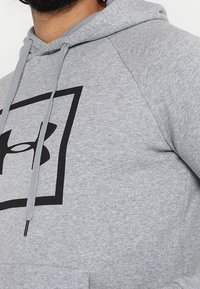 Sudadera gris con un logotipo negro dentro de un diseño cuadrado en la parte frontal. Tiene capucha con cordones y bolsillo delantero. Textura de tela suave.