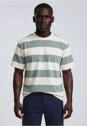 Homme à la peau foncée portant une chemise à manches courtes avec de larges rayures horizontales grises et blanches et un pantalon bleu foncé, debout sur un fond uni.