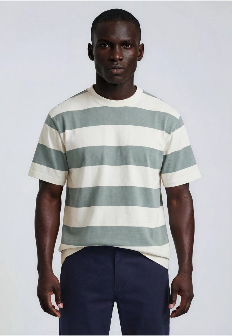 Homme à la peau foncée portant une chemise à manches courtes avec de larges rayures horizontales grises et blanches et un pantalon bleu foncé, debout sur un fond uni.