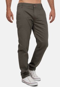 Pantalons en coton vert olive avec une coupe droite, présentant des accents de couture rouge et une fermeture à bouton. Portés avec des baskets blanches.