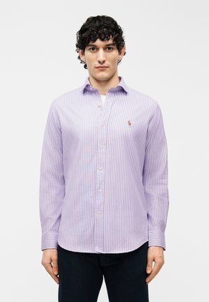 Mladý muž s kučeravými vlasmi, oblečený v svetlofialovej a bielo pruhovanej košeli na gombíky a tmavých nohaviciach, stojaci pred jednoduchým bielym pozadím.