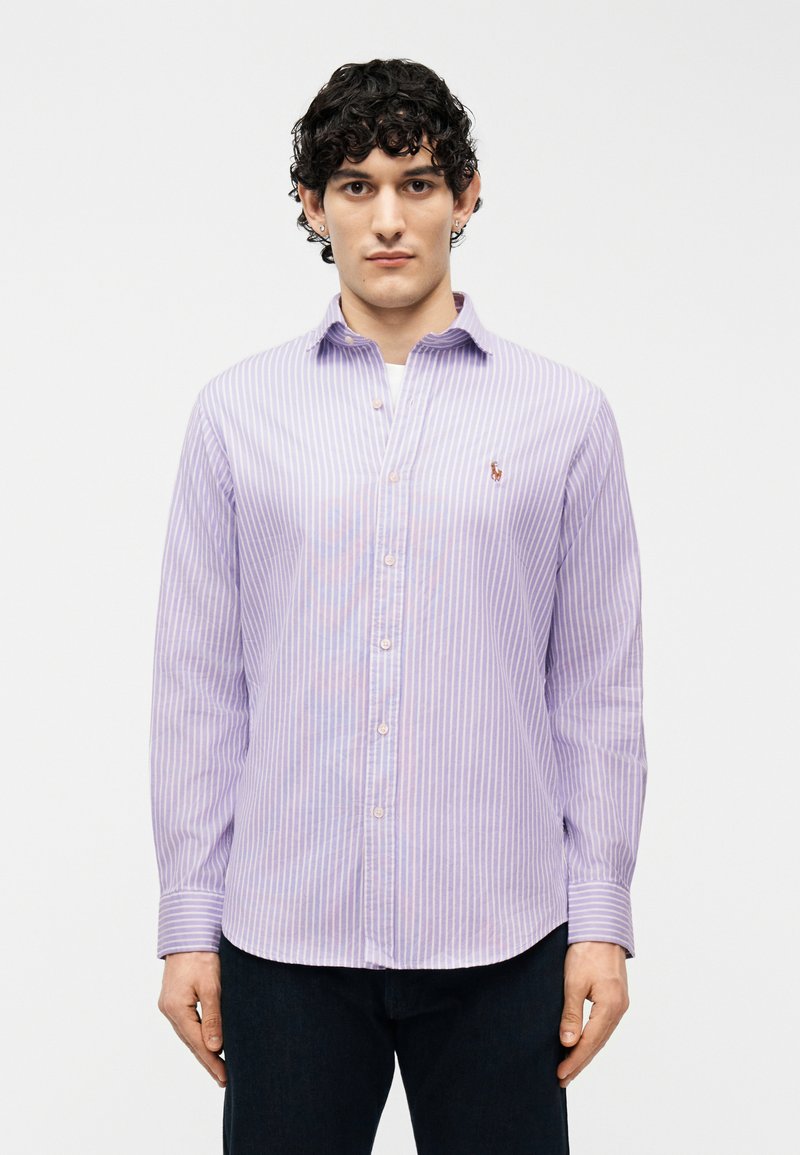 Νέος άνδρας με κατσαρά μαλλιά, που φοράει πουκάμισο με κατακόρυφες ροζ και λευκές ρίγες και σκούρο παντελόνι, στέκεται μπροστά σε λευκό φόντο.