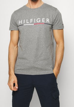 Mężczyzna ubrany w szarą koszulkę z krótkim rękawem z napisem "HILFIGER" i kolorowym paskiem, w połączeniu z ciemnoniebieskimi spodniami z boczną kieszenią.