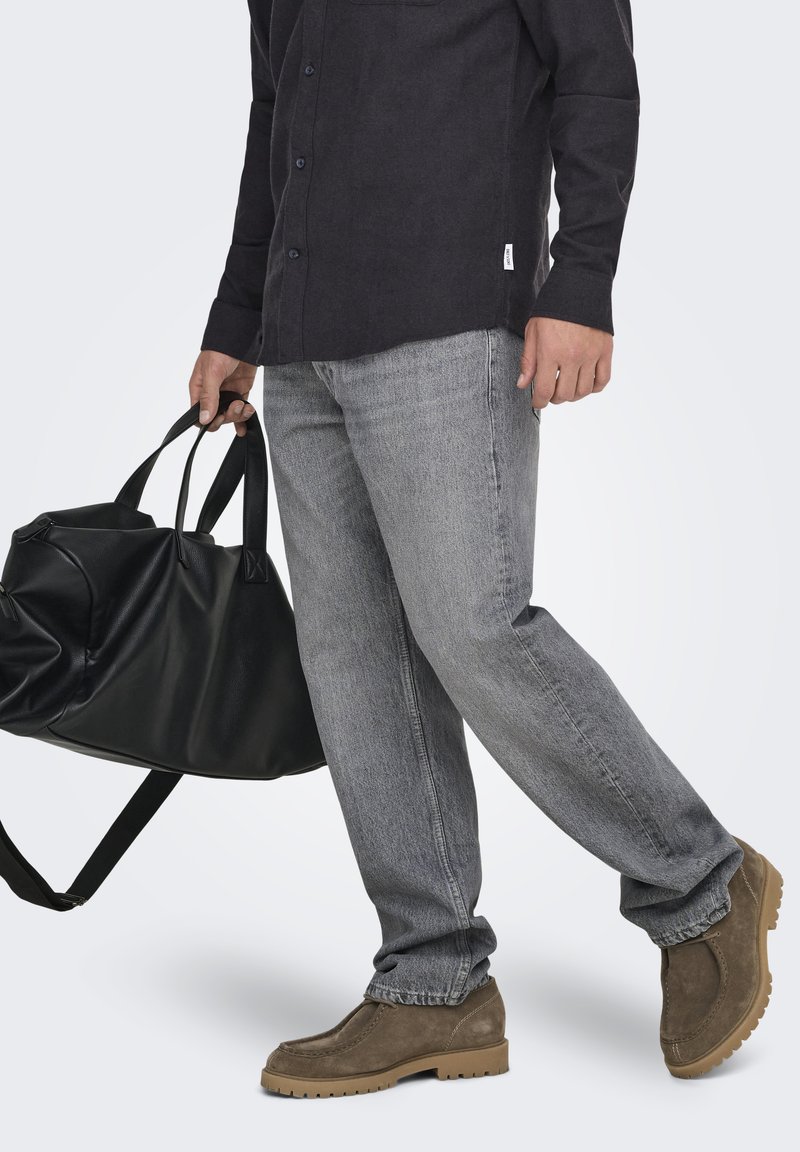 Homme portant un jean gris, des chaussures en daim marron et une chemise noire boutonnée, tenant un grand sac de voyage en cuir noir par les poignées.