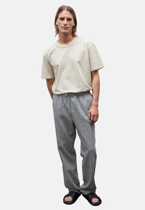 Homme portant un t-shirt beige uni, un pantalon rayé vertical noir et blanc, et des sandales noires, debout devant un fond uni.