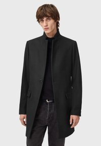 Mustat villasekoitekappa, jossa on korkea kaulus, yksittäinen nappisuljenta ja sivutaskut, päällä musta paita ja tummat farkut.