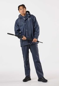 Marineblauw waterdicht jack met capuchon, drukknopen en trekkoorden, gecombineerd met bijpassende broek en zwarte schoenen; een zwarte paraplu vasthoudend.