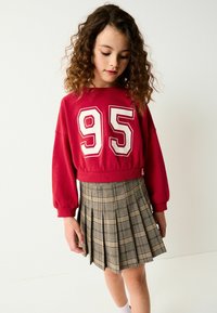 Rød cropped sweatshirt med stort hvidt "95" print, parret med en plisseret ternet nederdel i beige og sorte nuancer. Blødt stof og afslappet pasform.