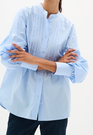 Osoba ubrana w jasnoniebieską plisowaną koszulę na guziki z długimi bufkami na rękawach, ze skrzyżowanymi ramionami, na jednolitym białym tle.