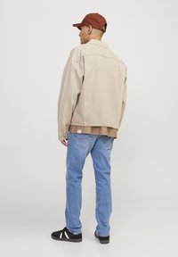 Chaqueta de mezclilla beige, jeans de un azul claro y zapatillas negras. Una gorra marrón realza el atuendo casual. Detalles de costura visibles en los jeans.
