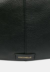 Borsa nera in pelle con superficie texturizzata, dettagli cuciti e un'etichetta dorata con la scritta "Coccinelle" in basso.