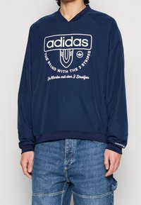 Mörkblå långärmad sweatshirt med v-ringning, med vit Adidas-logotyp och text. Strukturerad tyg med ribbade ärmslut och nederkant.