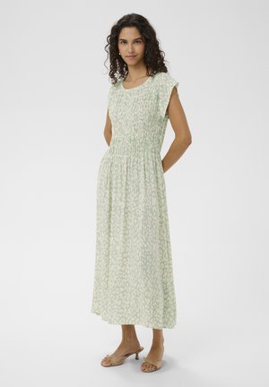 Donna in piedi con le mani dietro la schiena, indossa un vestito midi verde chiaro con motivo e sandali beige con tacco e punta aperta su sfondo bianco.