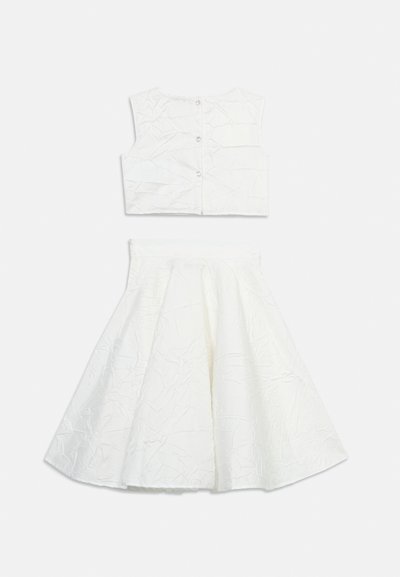 Λευκό αμάνικο κοντό τοπ με τρία κουμπιά μπροστά, σε συνδυασμό με φούστα μέχρι το μέσο της γάμπας, ψηλόμεση και με καμπάνα, και τα δύο με σατινέ υφή.