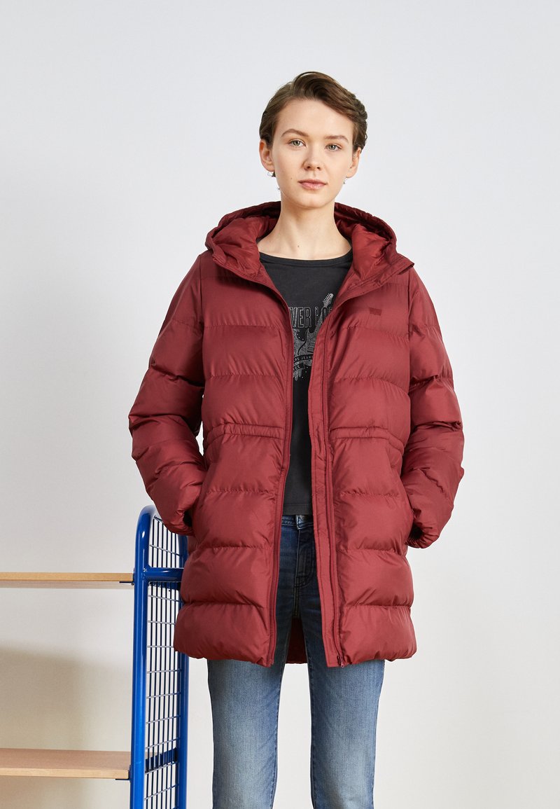 Giacca puffer bordeaux con cappuccio, caratterizzata da chiusura con zip, tasche frontali e un design trapuntato. Indossata sopra una maglietta scura e jeans.