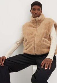 Weste aus Kunstfell in Tan mit hohem Kragen und Reißverschluss, mit seitlichen Taschen. Tragen über einem cremefarbenen Langarmoberteil und schwarzen weiten Hosen.