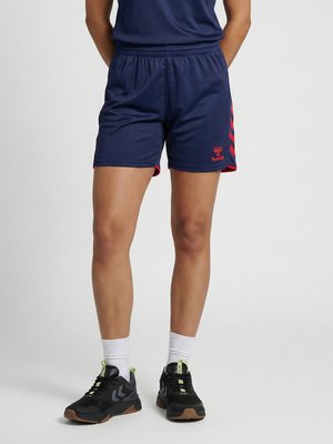 Person iført marineblå sportsshorts med røde accenter, hvide ankelsokker og sorte sportssko, der står mod en ensfarvet baggrund.