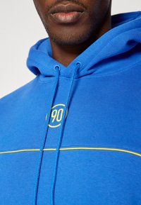Sudadera azul de mezcla de algodón con un bolsillo canguro. Presenta un logotipo amarillo bordado "90" y costuras de contraste a lo largo del pecho.
