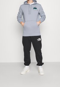 Sudadera gris con capucha y logo, con un bolsillo frontal y pantalones negros. Los pantalones tienen un logo de marca blanco. El modelo lleva zapatillas blancas.
