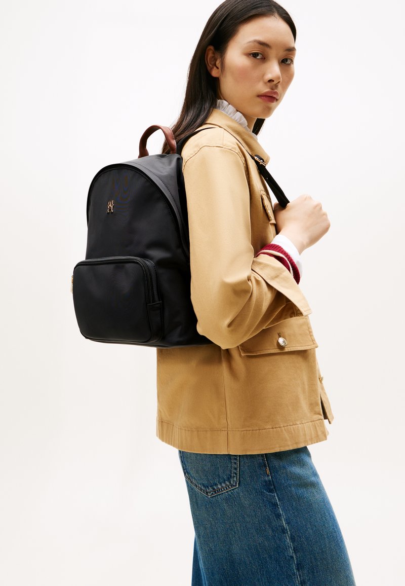 Woman wearing tan jacket and blue jeans carrying black backpack over one shoulder against plain white background.