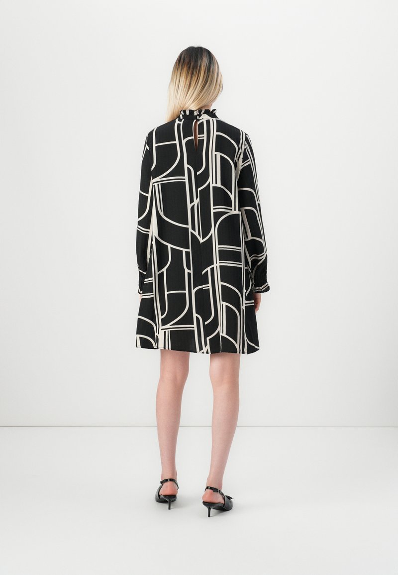 Mujer de pie, vistiendo un vestido de patrón geométrico en blanco y negro con mangas largas y zapatos de tacón bajo negros, de frente a un fondo blanco liso.