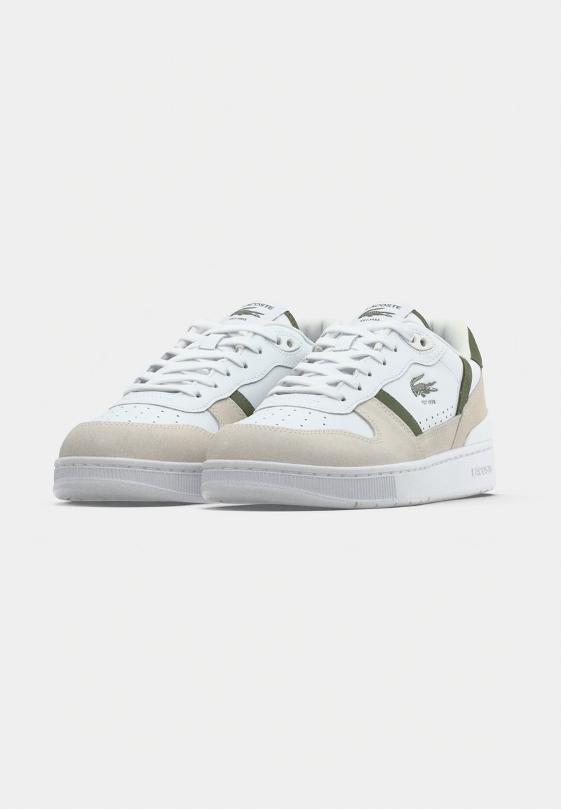 Baskets Lacoste blanches et beiges avec des accents verts et logo, design à lacets, bout perforé et semelle blanche sur un fond clair.