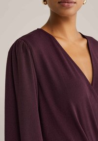 Donna che indossa un top a V di un profondo colore viola texture con dettaglio di spalle arricciato e orecchini a cerchio dorati, tagliati sotto il naso.