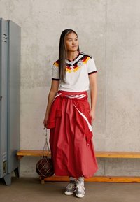 Λευκή αθλητική φανέλα με μαύρες, κόκκινες και κίτρινες λεπτομέρειες; κόκκινη φούστα δεμένη στη μέση; γκρι αθλητικά παπούτσια; γκρι σκηνικό από σκυρόδεμα; ξύλινος πάγκος.