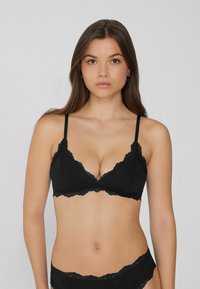 Femme aux longs cheveux bruns portant un soutien-gorge en dentelle noire et un sous-vêtement en dentelle noire assorti, sur un fond gris uni.