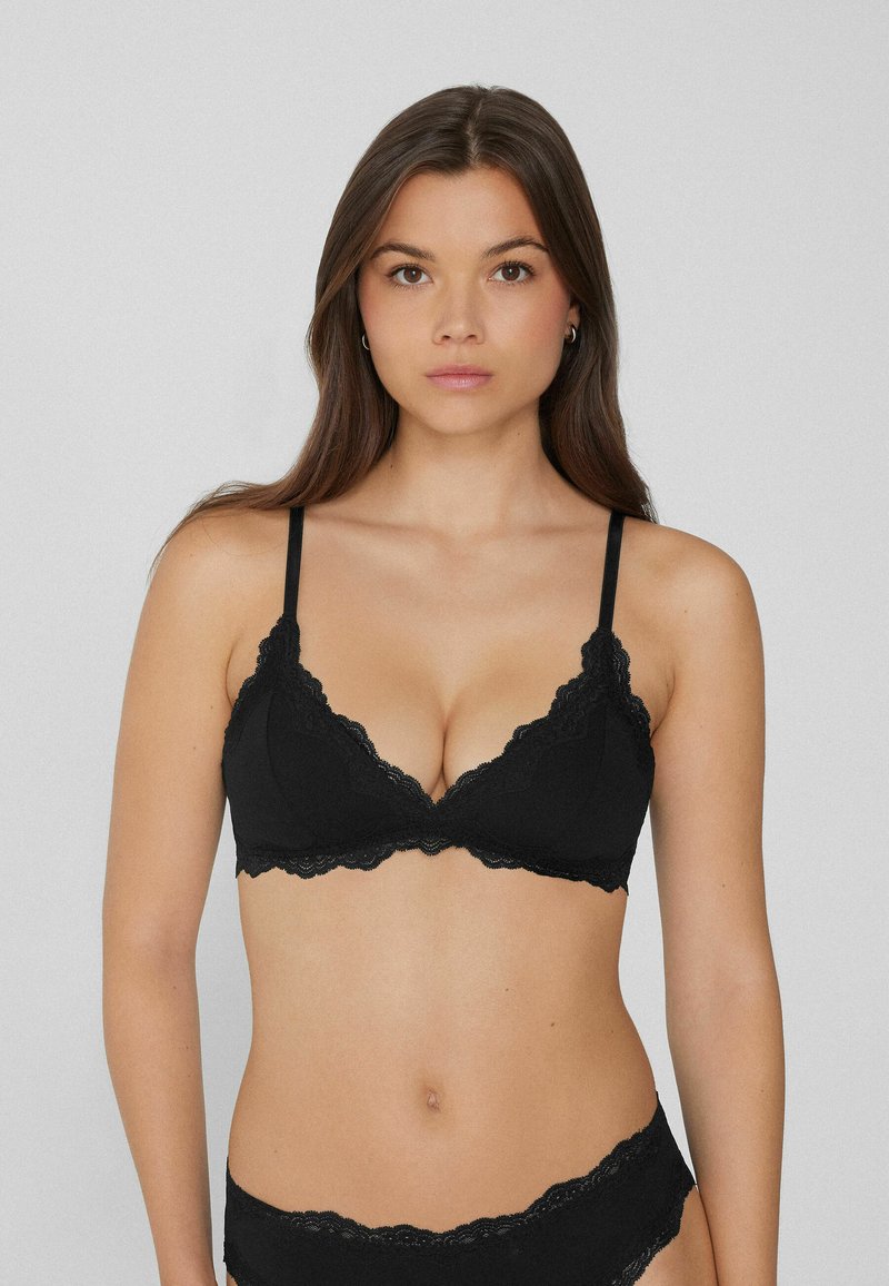 Femme aux longs cheveux bruns portant un soutien-gorge en dentelle noire et un sous-vêtement en dentelle noire assorti, sur un fond gris uni.
