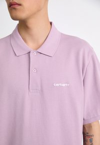 Polo shirt lilas clair avec une texture douce, deux boutons au col et un logo "Carhartt" blanc sur le côté gauche.