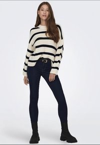 Strickpullover in Creme mit schwarzen Streifen, kombiniert mit dunklen Jeans und schwarzen Stiefeletten. Der Pullover hat einen lockeren Schnitt und einen weiten Ausschnitt.